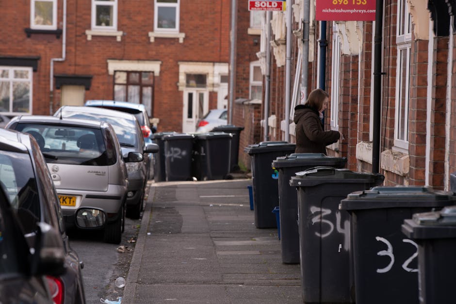 A narrow urban street scene featuring a sidewalk with several black wheelie bins lined up along the building facades, each with white spray-painted numbers. A woman dressed in a dark coat stands next to one of the bins, appearing to access its contents. To the left, parked cars occupy most of the parking spaces, with a silver hatchback closest to the foreground, showing visible rear lights and side mirrors. The background reveals brick terraced houses with white-framed windows and brickwork details, some with small front yards. The sky provides natural daylight, illuminating the scene evenly and highlighting the textures of the brickwork, the smooth plastic of the bins, and the metallic car surfaces. The environment suggests an everyday residential area where independent waste collection, such as bin lining and rubbish removal, takes place, with the presence of company-branded bins and markings indicating organized disposal or collection points.