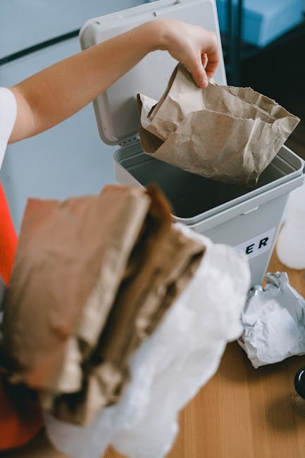 A person is seen disposing of paper waste into a small, light gray bin with a partially open lid. The individual’s hand is dropping a brown paper bag into the bin, which is positioned on a wooden surface. In the foreground, another crumpled brown paper bag is held in front of the camera, obscuring part of the view. The background includes a blue surface and a white object, possibly a piece of furniture or appliance, indicating an indoor setting. The scene illustrates common waste disposal activity, relevant to private rubbish collection or on-site clearance services for residential areas, with the focus on paper recyclables or general waste being prepared for collection. The overall environment appears clean and organized, emphasizing proper waste handling in accordance with domestic rubbish removal practices.