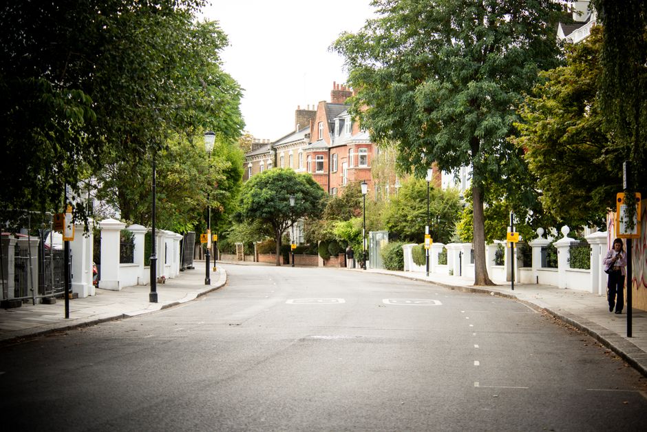 A quiet residential street in an urban area during daytime, featuring a gently curving asphalt road with white dashed lines marking parking bays. Flanking the street are white-painted low boundary walls with decorative finials and black wrought iron gates, which enclose front gardens of multi-storey Victorian-style houses constructed from red brick and stone, with bay windows and gabled roofs. Mature trees with dense green foliage line both sides of the street, their branches extending over the pavement and road, providing shade and a natural canopy. Along the pavements, there are regularly spaced black lampposts with white globular lights, and some yellow and black street bollards, possibly indicating parking restrictions or pedestrian zones. On the right sidewalk, a person is walking while carrying a bag, and on the left side, a bicycle is locked to a railing near the house entrance. The scene appears to be part of an established neighbourhood that may require private rubbish collection, with [COMPANY_NAME] providing an alternative waste disposal service to local authority options, supporting efficient rubbish removal and street cleanliness management.