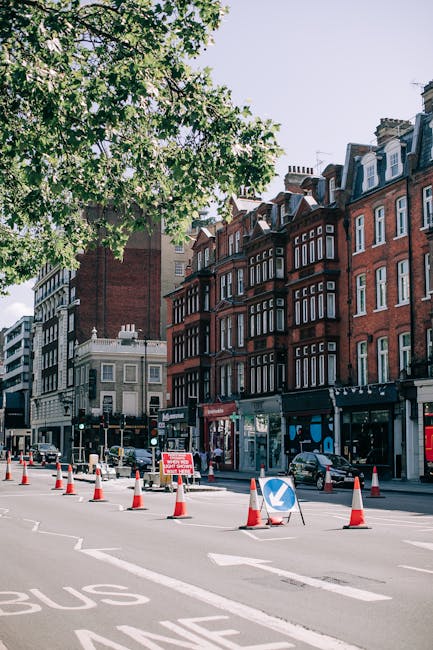 A wide street scene in an urban area shows a row of multi-story red brick buildings with white-framed windows and roof details lining the right side of the image. The pavement in front of the buildings hosts several parked cars, and a section of roadway is marked with traffic cones and a directional blue arrow sign, indicating ongoing traffic management or rerouting. A pedestrian crossing is visible with a black and white striped striped signal box, while the street includes a dedicated bus lane marked with partial white lettering. On the left side of the image, a large leafy tree with vibrant green foliage extends branches across the top, casting partial shade over the scene. The area appears busy with some additional vehicles and street signage visible in the background. The overall atmosphere suggests an active urban environment, where managing waste or rubbish removal could involve nearby services or collection points, fitting with practices for private waste handling and alternative rubbish disposal options. The clear daylight and cityscape atmosphere contribute to an accurate, neutral representation of this street scene for accessibility and SEO purposes related to rubbish and waste removal services in urban settings.