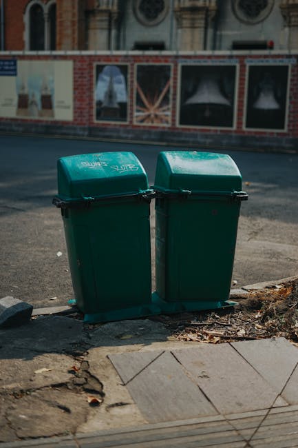 Two green wheeled rubbish bins with hinged lids positioned on a sidewalk curb at the edge of a paved street, with visible black handle mechanisms on the lids. The bins are made of plastic with a textured surface and slightly rounded tops, showing signs of outdoor weather exposure. They are placed on a concrete curb next to a footpath, with some fallen leaves and debris around their base. The background features a building with brick and plaster exterior, large windows, and decorative architectural details, as well as a road surface with some markings. The scene is illuminated by natural daylight, indicating an outdoor urban setting suitable for private waste collection services like those offered by rubbish removal companies in the area, supporting alternative waste handling options outside local authority collection.