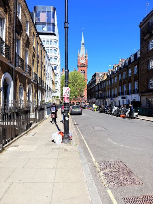 A wide city street scene featuring a clear blue sky above rows of Victorian brick terraced buildings with black wrought iron railings and white window frames on the left side. The pavement is clean and has a few pedestrians; one person is walking past a white plastic bag of rubbish placed next to a black lamppost which also displays a warning sign. On the right side, there are parked motorcycles and a small black car near the curb. In the background, a prominent red brick church with a tall, pointed spire and clock face is visible, surrounded by modern high-rise buildings that include a glass-fronted tower with curved structural elements. The scene captures a typical urban environment where private rubbish collection services like [COMPANY_NAME] might carry out on-site clearance or alternative waste handling, with visible waste or rubbish accumulation on the sidewalk in the form of the bag and some loose items, conveying a context relevant to rubbish removal near central London landmarks.