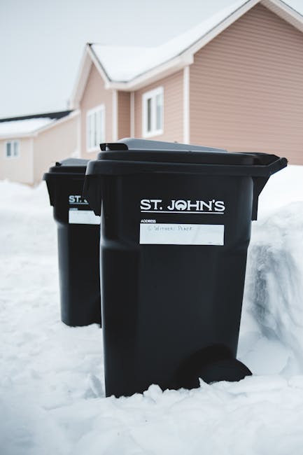 Two black plastic wheelie bins with white labels, positioned outside on a snow-covered pavement in front of a residential building with light beige siding and white-framed windows. The bins are marked with the label 'ST. JOHN'S,' indicating private waste collection. The scene is set during winter, with snow accumulating around the bases of the bins and partially covering the surrounding ground. The bins are closed and appear to be used for rubbish disposal, possibly for an independent waste removal service. The background features the side of a modern, two-story house with a pitched roof, and the environment suggests a suburban neighbourhood. The image reflects the outdoor storage of waste receptacles awaiting collection, aligning with non-local authority rubbish management methods and alternative waste handling options. The overall lighting is natural, with soft daylight illuminating the scene, emphasizing the texture of the snow and the matte finish of the trash bins.